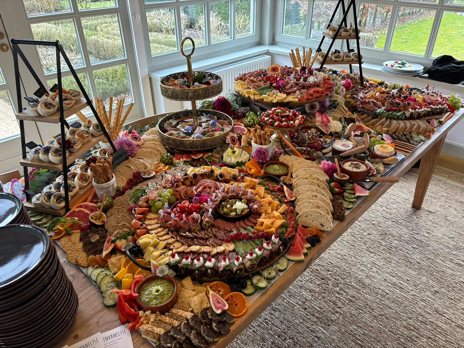 Lange grazing table opgebouwd in een serre, met etagères, broodjes en verse bloemen